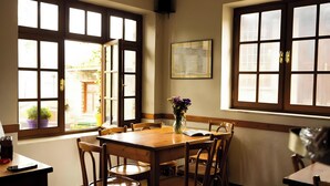 A dining area with a wooden table and chairs, large windows with multiple panes, and a vase with flowers on the table. - Guest House 'Armoloi Room 2' with Mountain View, Shared Terrace and Wi-Fi (Konitsa)
