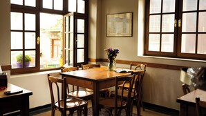 Un comedor con mesa y sillas de madera, ventanas grandes con múltiples divisiones y un jarrón con flores sobre la mesa.