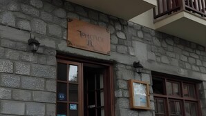 A stone building with a signboard, wooden balcony railings, and a table with chairs outside.