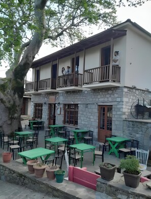 A two-story building with a stone facade, outdoor seating with green tables and chairs, and a balcony with two people. - Guest House 'Armoloi Room 5' with Mountain View, Shared Terrace and Wi-Fi (Konitsa)