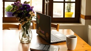 A wooden table with a laptop, a cup of coffee, and a vase of flowers by a window with multiple panes.