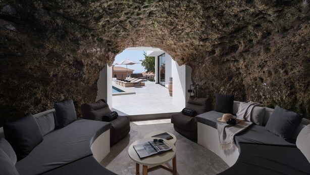 Un intérieur moderne avec un espace salon circulaire, une petite table et une vue sur un espace extérieur accessible par une arche naturelle en roche, cet espace extérieur comprenant une piscine et un coin détente.