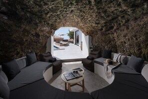 Un intérieur moderne avec un espace salon circulaire, une petite table et une vue sur un espace extérieur accessible par une arche naturelle en roche, cet espace extérieur comprenant une piscine et un coin détente.