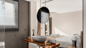 Une salle de bain moderne avec un grand lavabo sur pied, un meuble-lavabo en bois, un miroir et un lit recouvert d’une couette blanche.