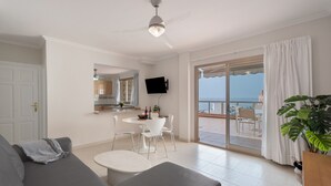 Una sala de estar moderna con un sofá gris, una mesa de comedor con sillas, un televisor de pantalla plana en la pared y un balcón con vista al mar.