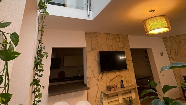 A modern interior with a high ceiling, a TV mounted on a stone wall, and a staircase leading to an upper level. - Rural House 'La Morena' with Mountain Views, Wi-Fi, and Air Conditioning (Guaro)