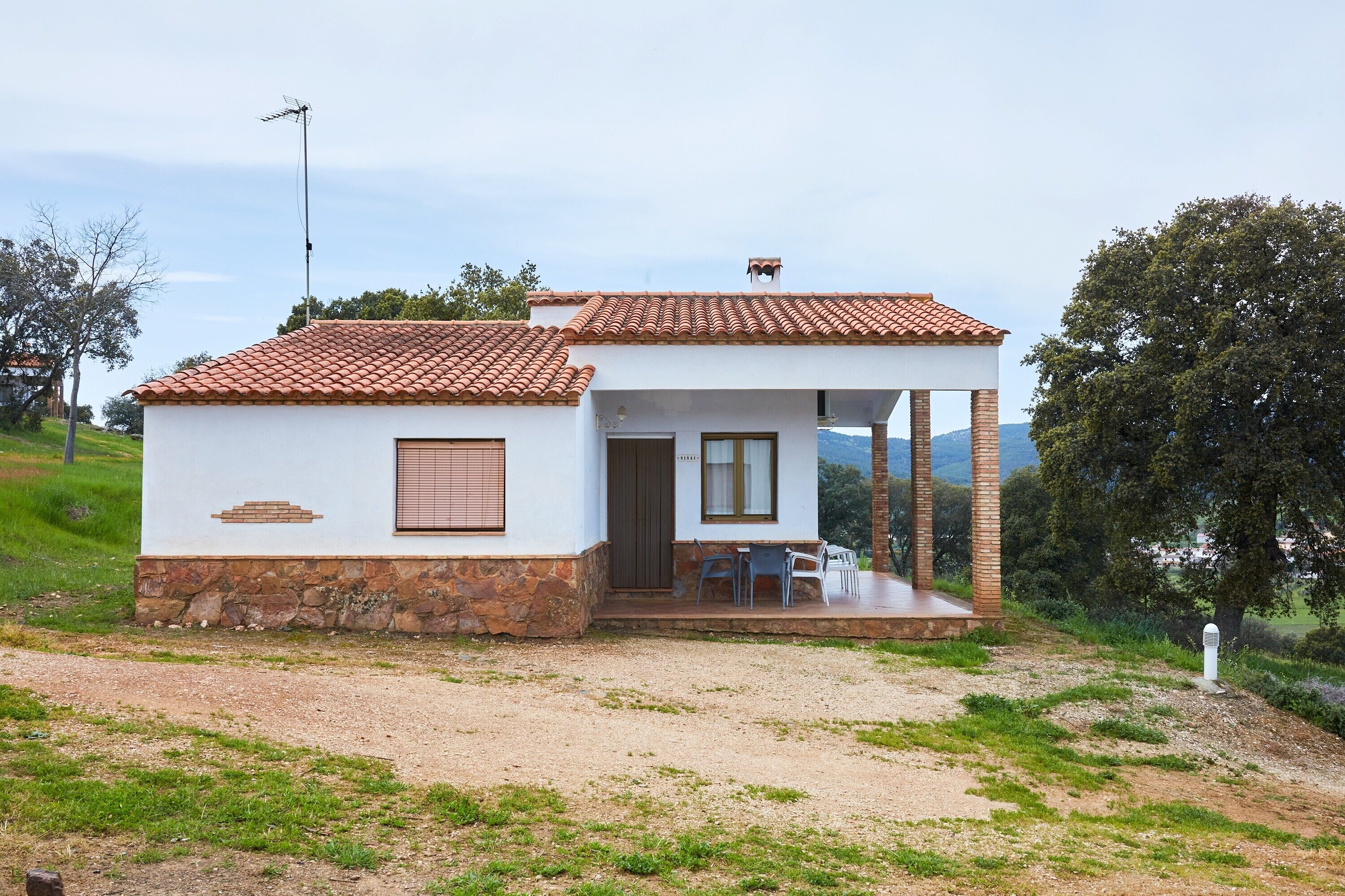 A small, single-story house with a tiled roof, a porch with chairs, and a stone foundation.