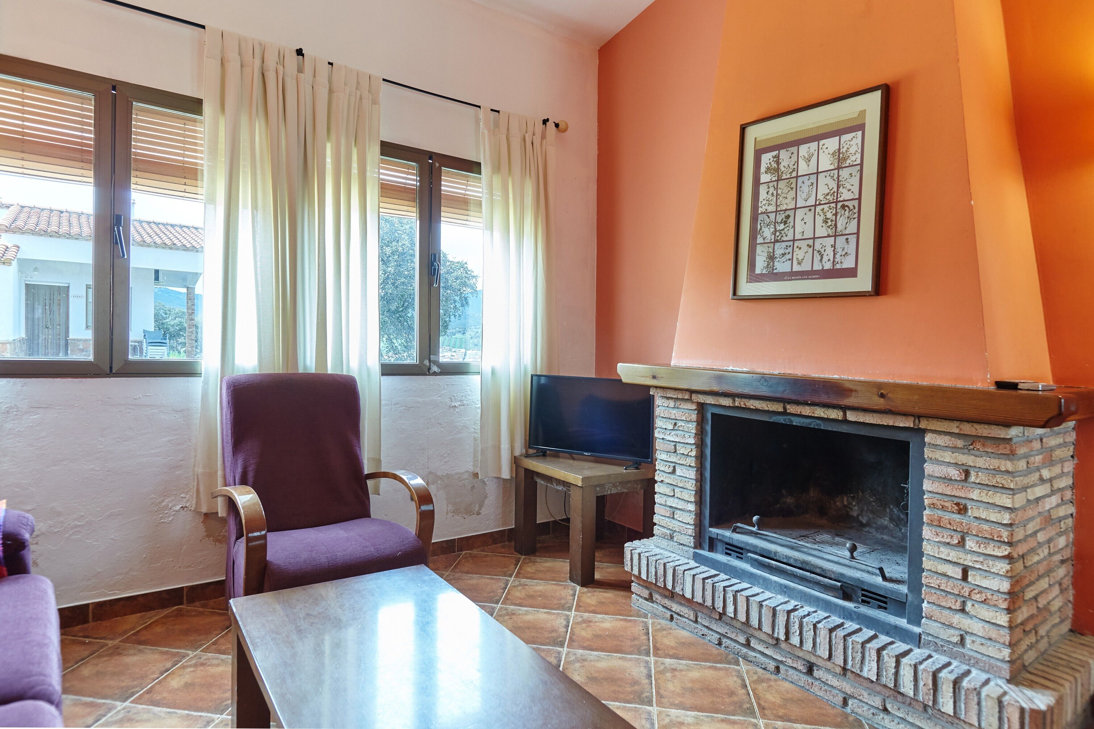 Un salon avec une cheminée en briques, un téléviseur posé sur une petite table, un fauteuil violet et une fenêtre avec des rideaux.