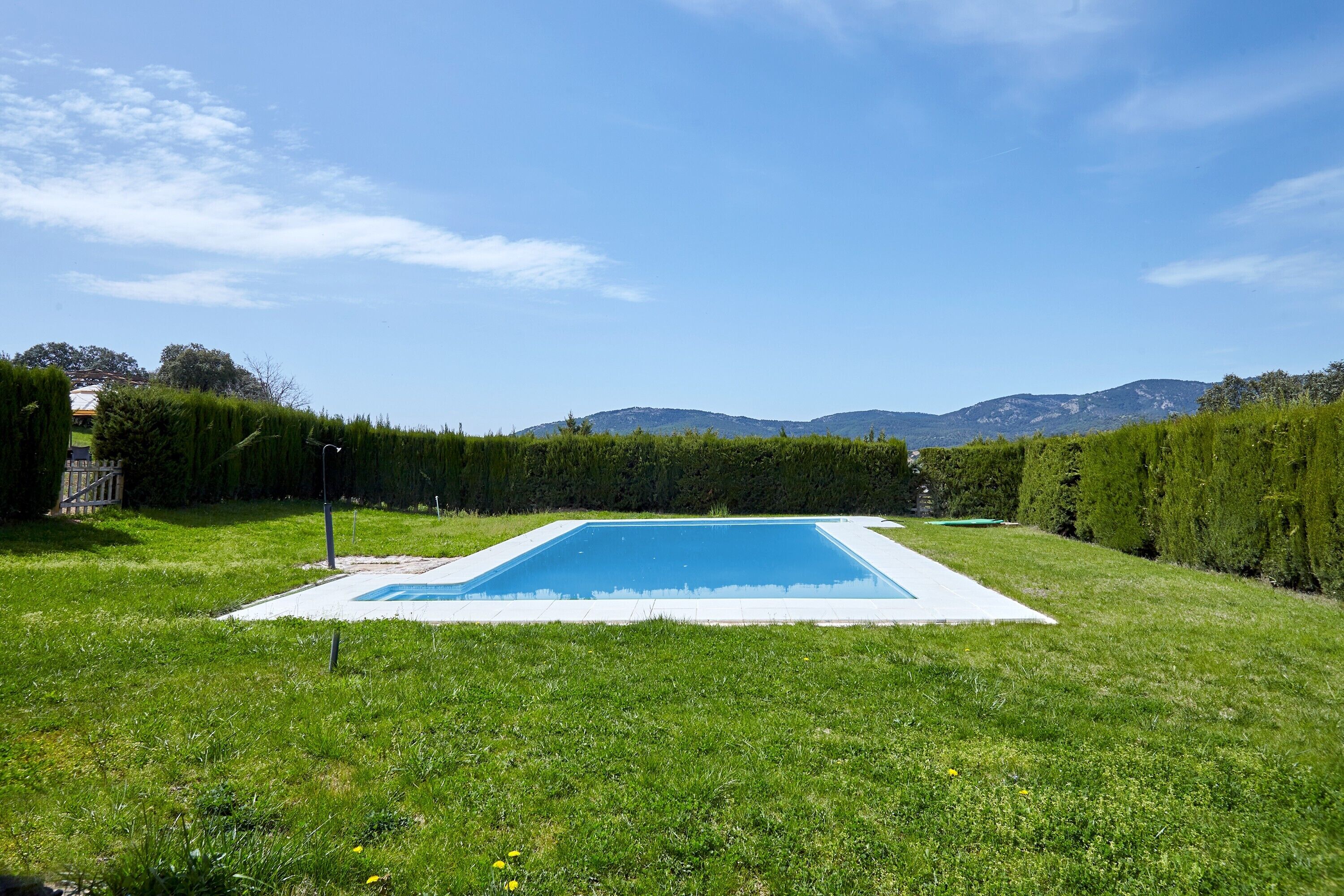 Una piscina rectangular con borde blanco, rodeada de césped y setos bien cuidados, y montañas al fondo.