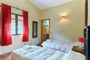 A bedroom with two beds, a window with red curtains, a wooden door, and a wall-mounted lamp.