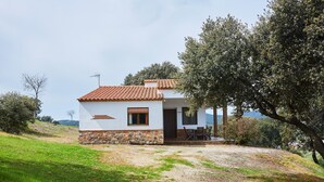 Un pequeño edificio blanco con techo de tejas, base de piedra y una puerta que da a un porche cubierto con asientos al aire libre.