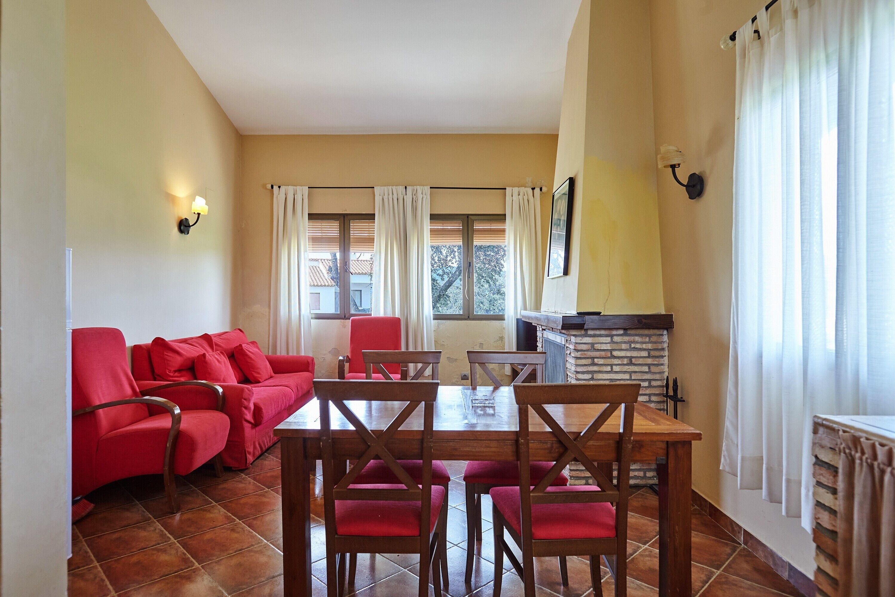 Una sala de estar con un sofá rojo, una mesa de comedor de madera y sillas, una chimenea y ventanas grandes con cortinas.