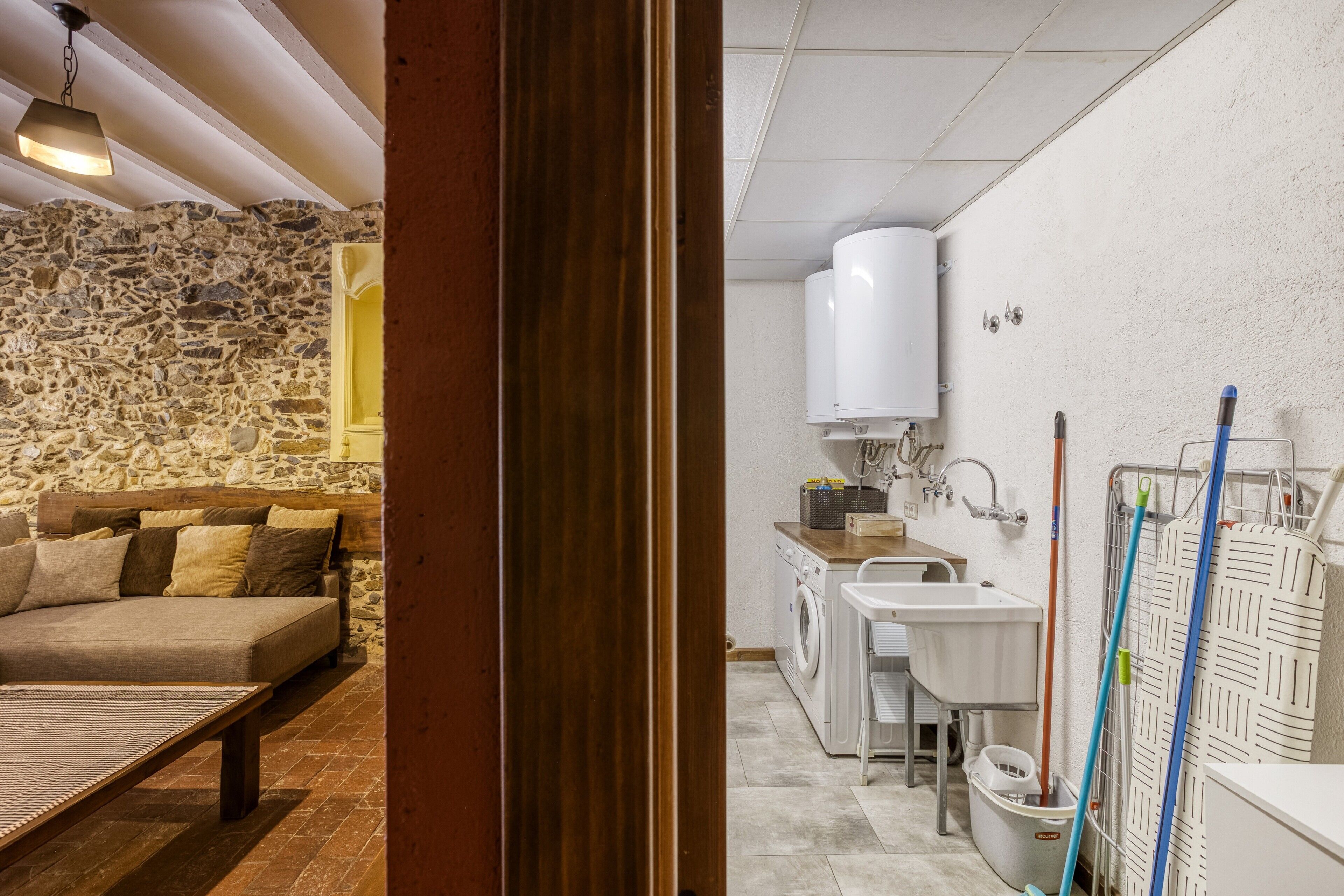 A room with a stone wall, a sofa, a table, a washing machine, and a sink.