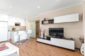 Een moderne keuken met witte kasten, een magnetron en een spoelbak. Er is een eethoek met een witte tafel en stoelen. Een flatscreen televisie is aan de muur bevestigd boven een donker houten console.