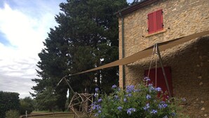 Ein Steinhaus mit roten Fensterläden, einer Schaukel und einem Garten mit violetten Blumen.