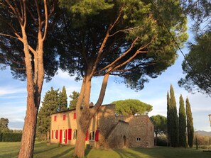 Un bâtiment en pierre avec des volets rouges, entouré d’arbres et d’une zone herbeuse.