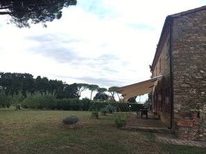 Edificio de piedra con un patio cubierto, mobiliario exterior y un jardín con césped, árboles y arbustos.