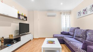 A modern living room with a flat-screen TV, a white coffee table, a purple sofa, and a white cabinet with shelves.