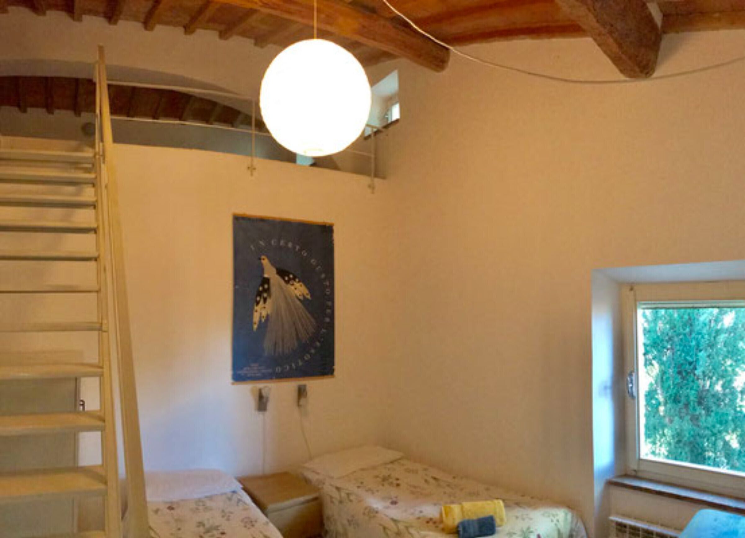 Ein Zimmer mit einer Holztreppe, einem Bett mit floralem Überwurf, einem Poster an der Wand und einem Fenster mit Blick auf Bäume.