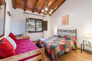 Une chambre à coucher avec un lit en bois, une couverture colorée, une fenêtre avec des rideaux, une lampe fixée au mur et un plafond avec des poutres apparentes.