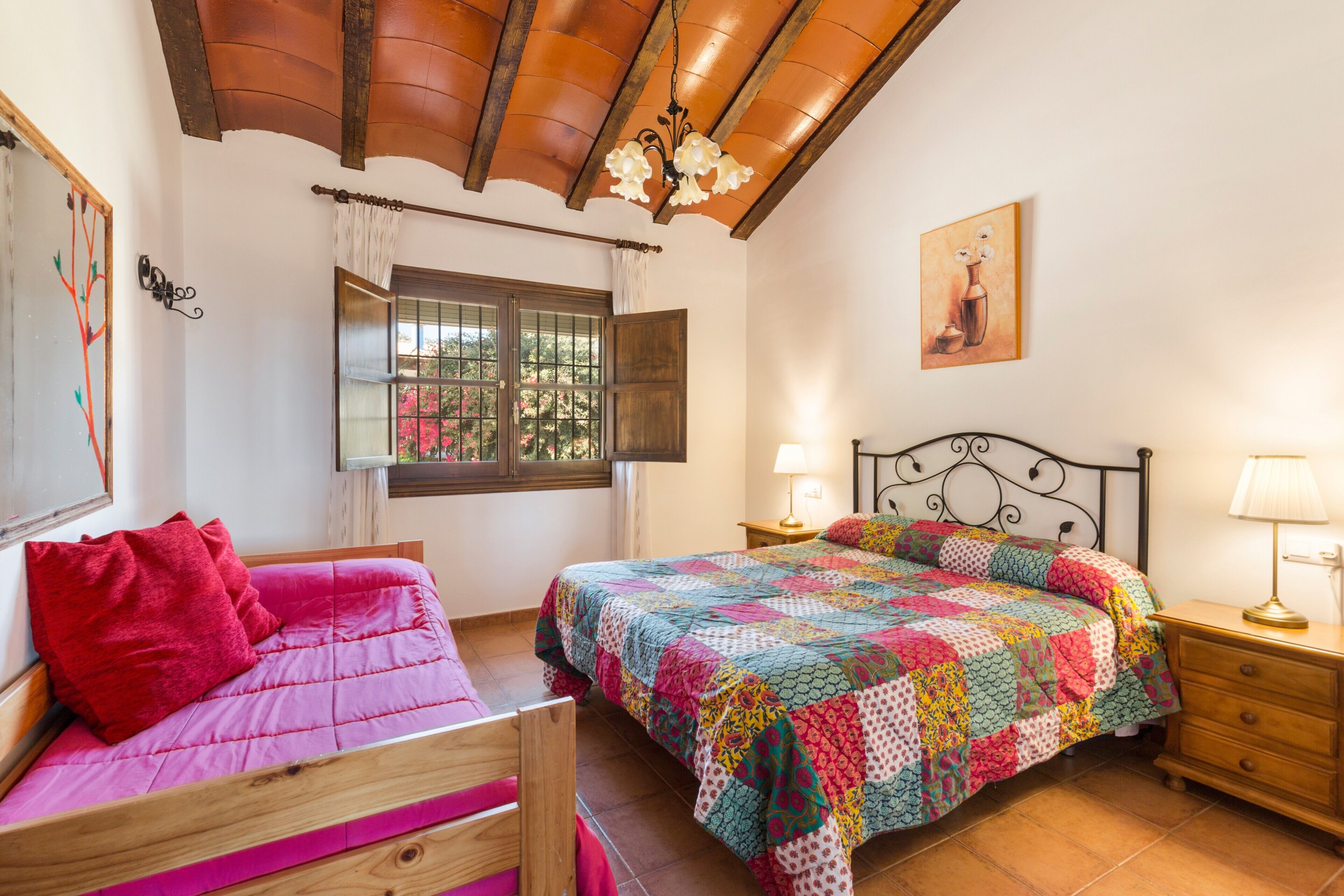 Un dormitorio con una cama de madera, una cama individual, una mesita de noche con lámpara, una ventana con cortinas y un cuadro en la pared.