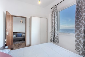 Une chambre à coucher comprenant un lit, une table de chevet, une armoire, un miroir, une fenêtre avec des rideaux et une vue sur la mer.