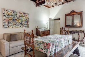 Une pièce comprenant un canapé, une table recouverte d’une nappe à motifs floraux, une chaise en bois, un meuble et un grand tableau abstrait accroché au mur.