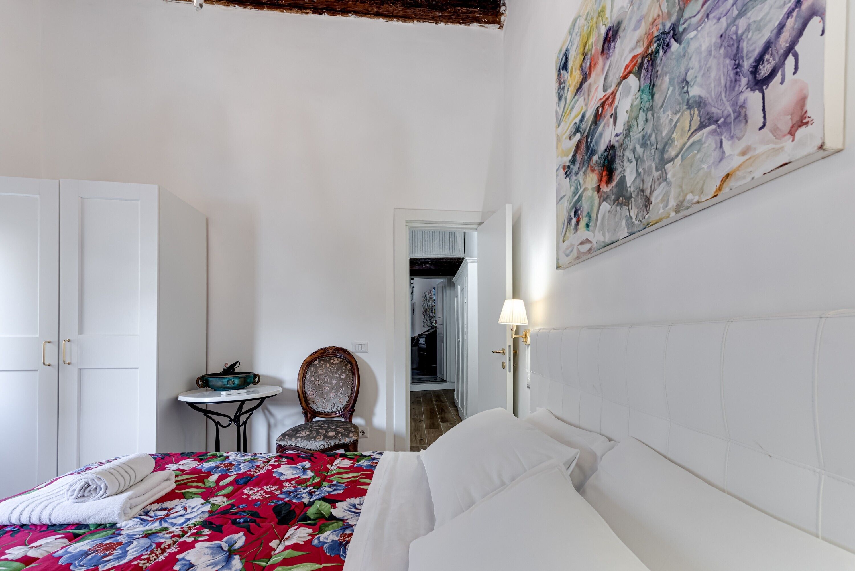A bedroom with a bed, a chair, a table, a painting, and a door leading to another room.