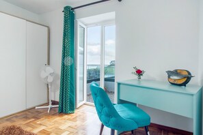 A room with a teal chair, a white fan, a desk with a bird sculpture, and a view through a glass door.