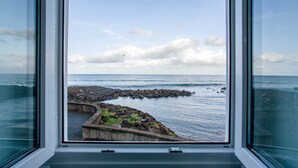 A view through a window of a coastal area with a rocky breakwater and calm sea. - Apartment 'Beach Front Flat' with Sea View, Private Terrace and Wi-Fi (São Vicente)