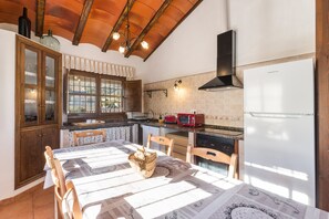 A kitchen with wooden cabinets, a dining table, chairs, a refrigerator, and a window with curtains.