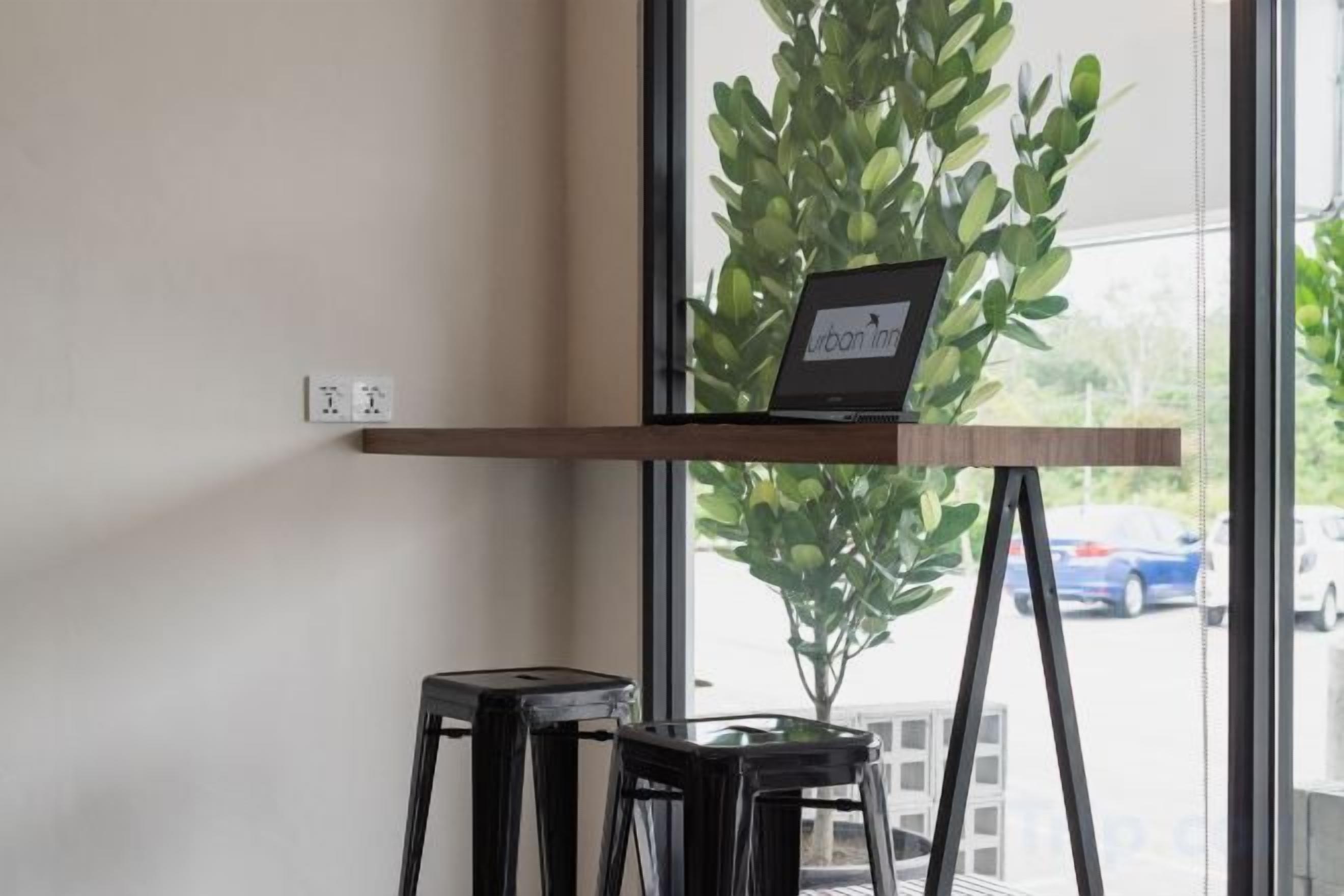 A laptop on a wooden shelf, a potted plant, and two black stools in a room with a window.