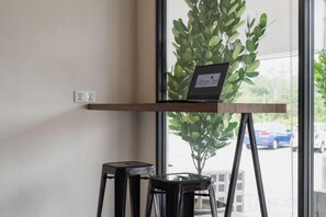 A laptop on a wooden shelf, a potted plant, and two black stools in a room with a window. - Urban Inn, SP Saujana (Sungai Petani)