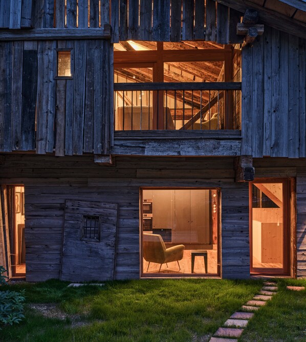 A rustic wooden building with a staircase, large windows, and a grassy yard.