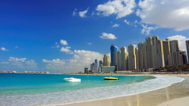 A beach with clear turquoise waters, a sandy shore, and a city skyline with high-rise buildings in the background. - 2BR apartment Sea View Upgraded on JBR (Dubai)
