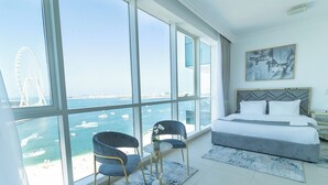 Een moderne hotelkamer met een groot raam dat uitzicht geeft op zee en een brug. Er staat een bed met een grijze hoofdbord, een nachtkastje en twee stoelen.