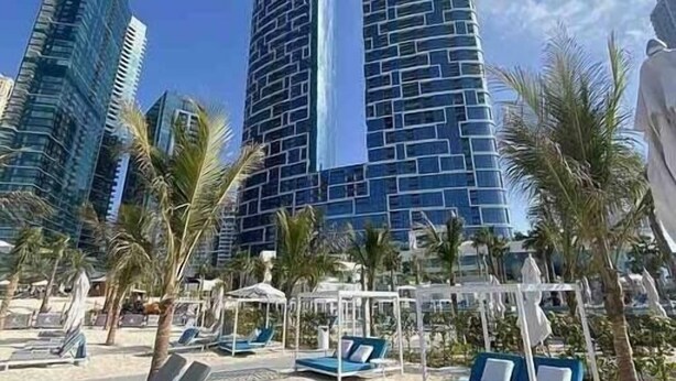 Una platja amb gandules i palmeres, un edifici modern amb una façana única, i altres edificis alts al fons.