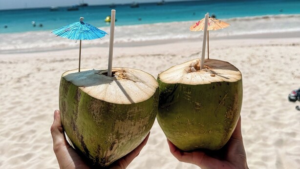 Two hands holding coconut drinks with straws, one with a blue umbrella and one with a straw, on a beach with a clear sky and calm sea in the background. - Heliconia Room Eden On The Sea Carlisle Bay Barbados  (Bridgetown)
