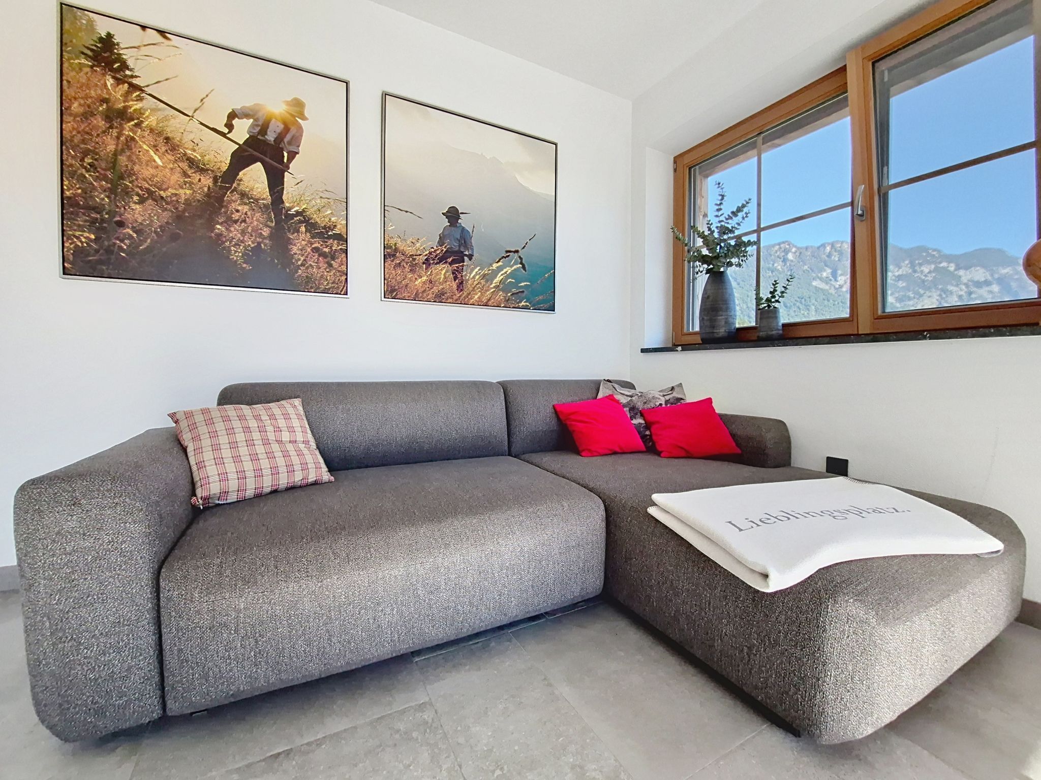 Een moderne woonkamer met een grijze modulaire bank, een rode en een roze kussen, en een witte plaid. Boven de bank hangen twee ingelijste foto's.