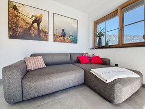 A modern living room with a grey sectional sofa, a red and a pink pillow, and a white throw blanket. There are two framed pictures on the wall above the sofa.
