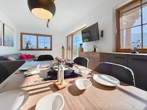 A dining area with a table set for four, a flat-screen TV mounted on the wall, and a window with a view of mountains.