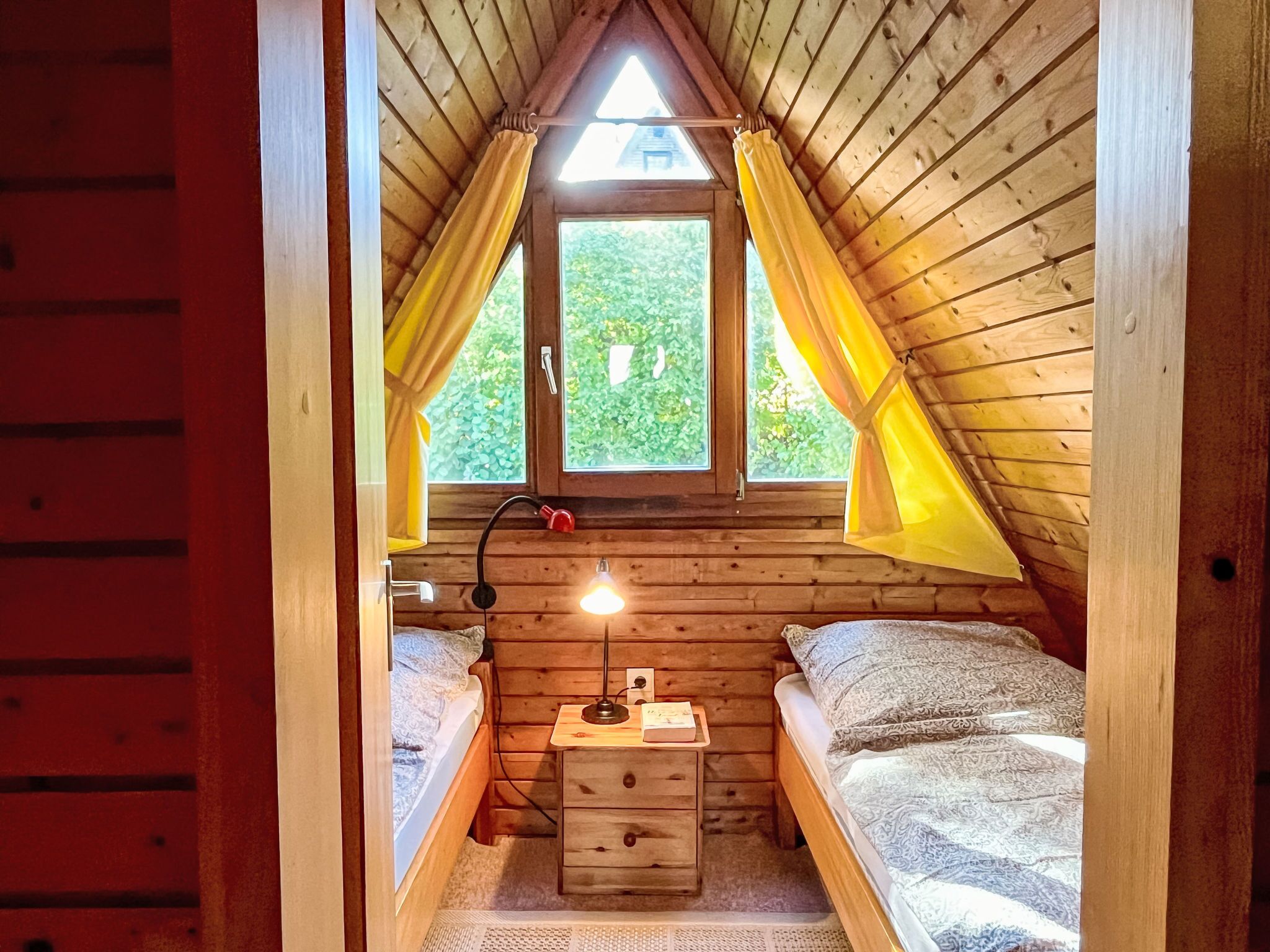Ein gemütliches Schlafzimmer im Dachgeschoss mit schrägen Wänden, Holzboden, zwei Betten, einem Nachttisch, einer Lampe und einem Fenster mit Vorhängen.