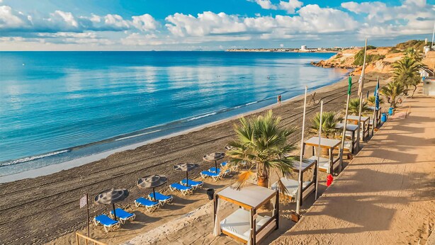 Una playa con una fila de tumbonas y sombrillas, un paseo marítimo de arena y vistas al mar.