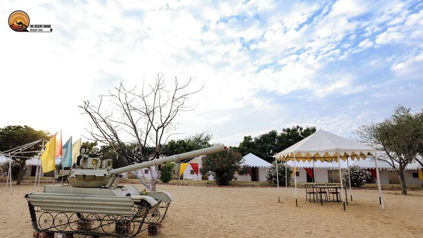 A military tank on display in a sandy area with tents and flags in the background. - The Desert Dhani Resort (Jaisalmer)