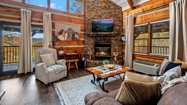 Una sala de estar acogedora con chimenea de piedra, un sofá confortable, una mesa de centro de madera y ventanas amplias con vistas al exterior.