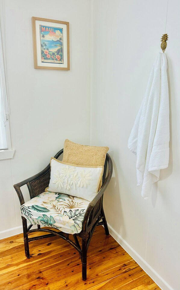 A room with a wooden floor, a wicker chair with a floral cushion, a framed picture on the wall, and a white towel hanging on a hook. - Hali'a - Renovated, 3 Bedroom Beach Cottage in Burleigh 5 mins walk to beach (Burleigh Heads)