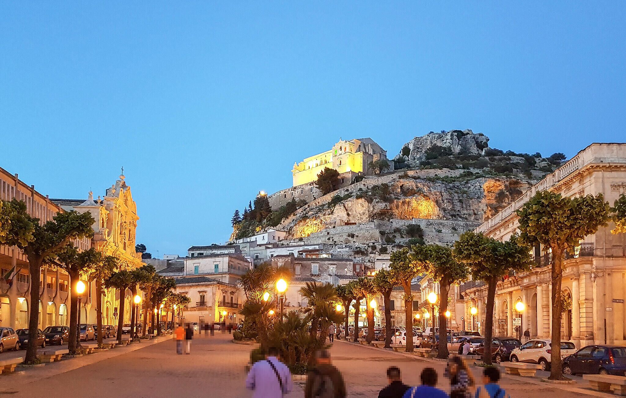 A historic city square with a prominent building on a hill, street lamps, and parked cars.