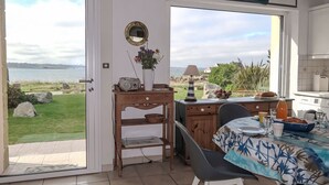 Ein Essbereich mit Meerblick, ein Holztisch mit Tischdecke, ein Stuhl, ein Radio und eine Vase mit Blumen.