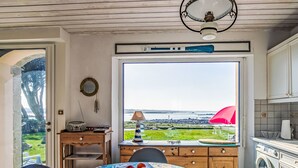 Ein Essbereich mit Tisch für zwei, Blick auf das Meer durch ein großes Fenster und eine Waschmaschine im Hintergrund.