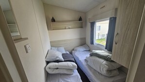 A compact bedroom with two single beds, a small shelf with decorative items, a window with curtains, and a power outlet on the wall. - Ruby Cabin Sleeps 6 with Free Parking (Rhyl)
